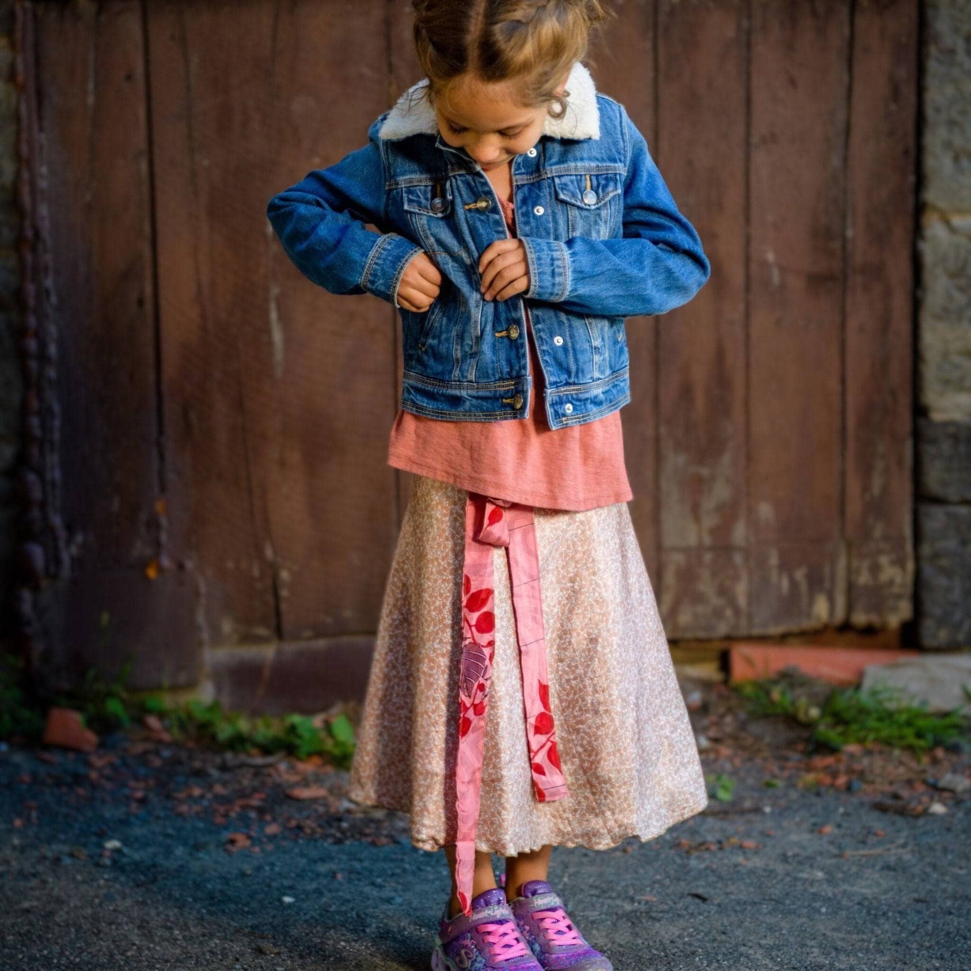 Kid model is standing outside in front of a wooden door wearing a light pink mini sari wrap skirt. 
