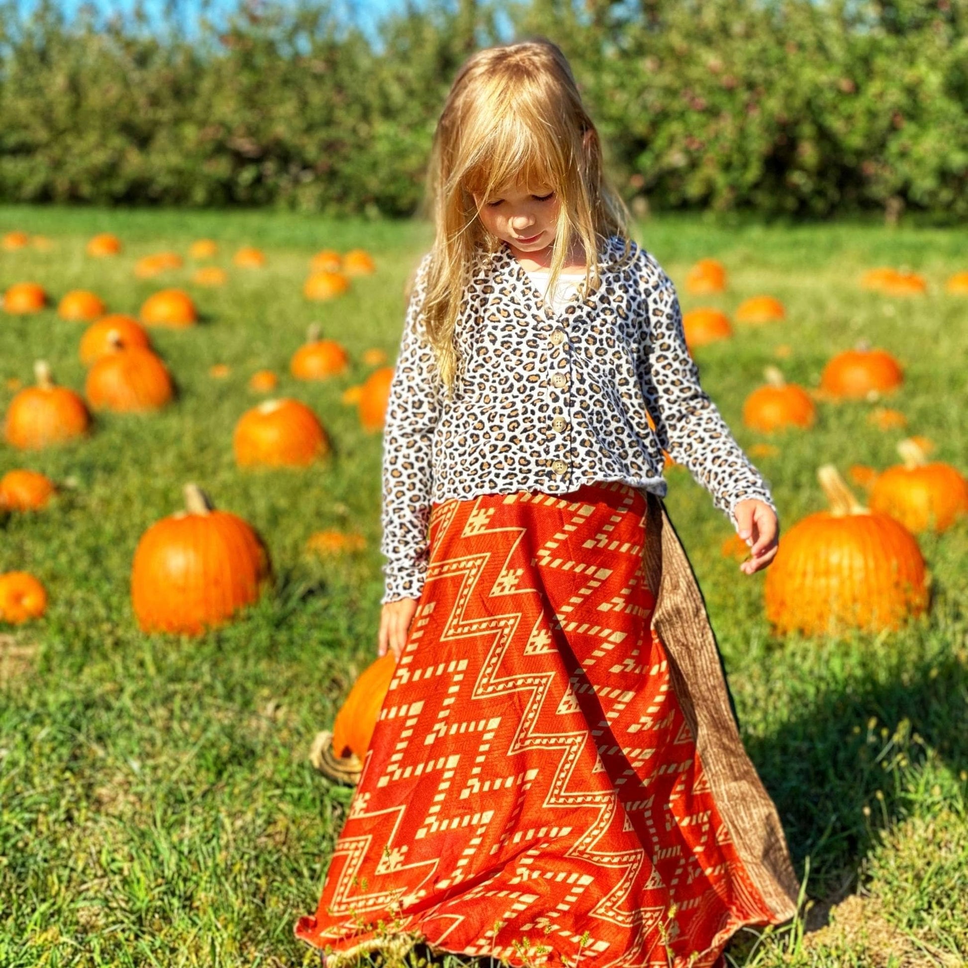 Kid model walking through a field of pumpkins wearing a  maxi sari wrap skirt in orange. 