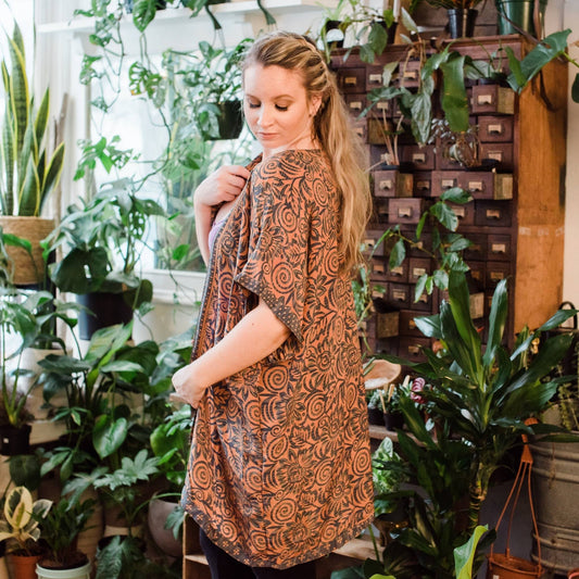 Model is looking over shoulder wearing an orange amara short duster while standing in front of some potted plants.