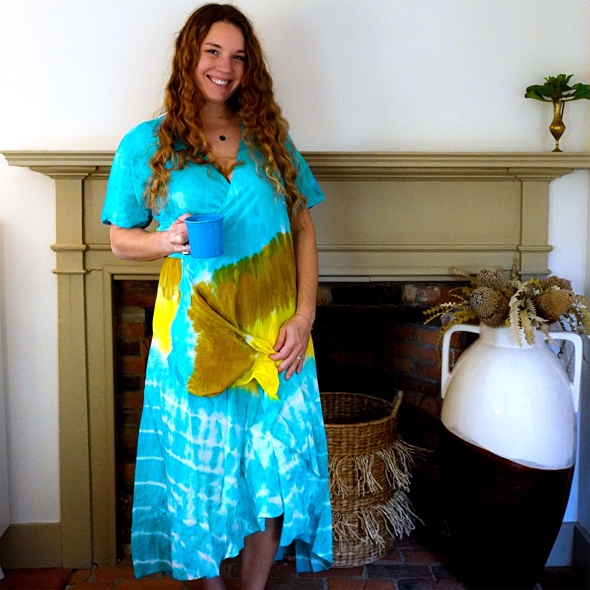 Woman standing in front of fireplace with blue coffee mug and tie dye wrap dress smiling at the camera