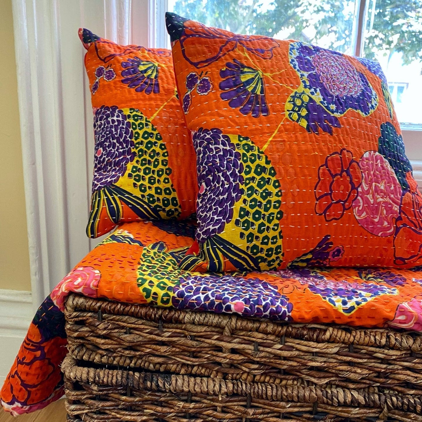 Two orange symphony kantha pillows laying a a matching quilt on top of a wooden basket in front of a window.
