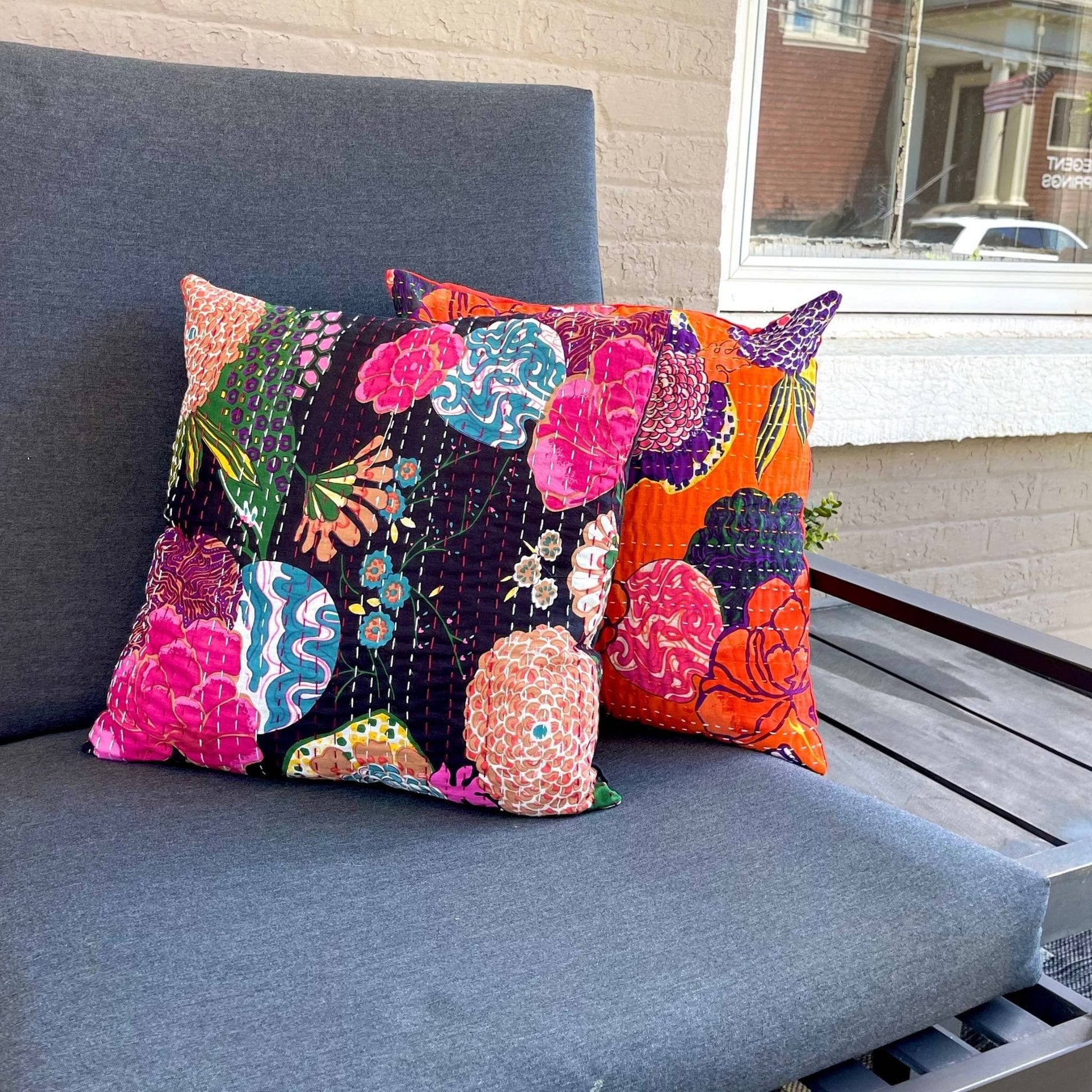 Two kantha pillows (in colors night lily and orange symphony) sitting on a grey couch outside.  