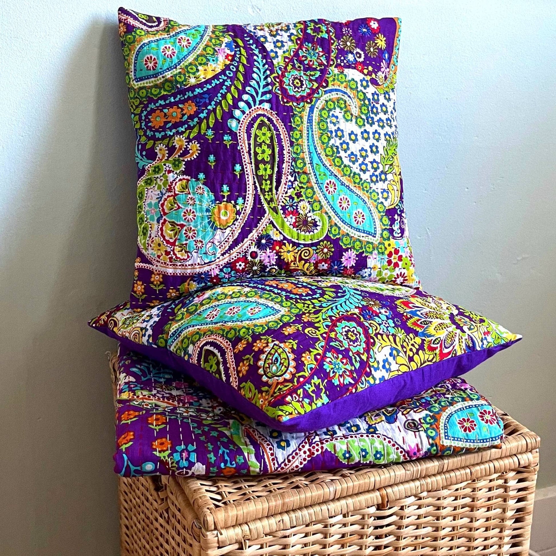 Two purple kantha pillow covers sitting on a matching quilt on a wooden hamper in front of a light blue wall.