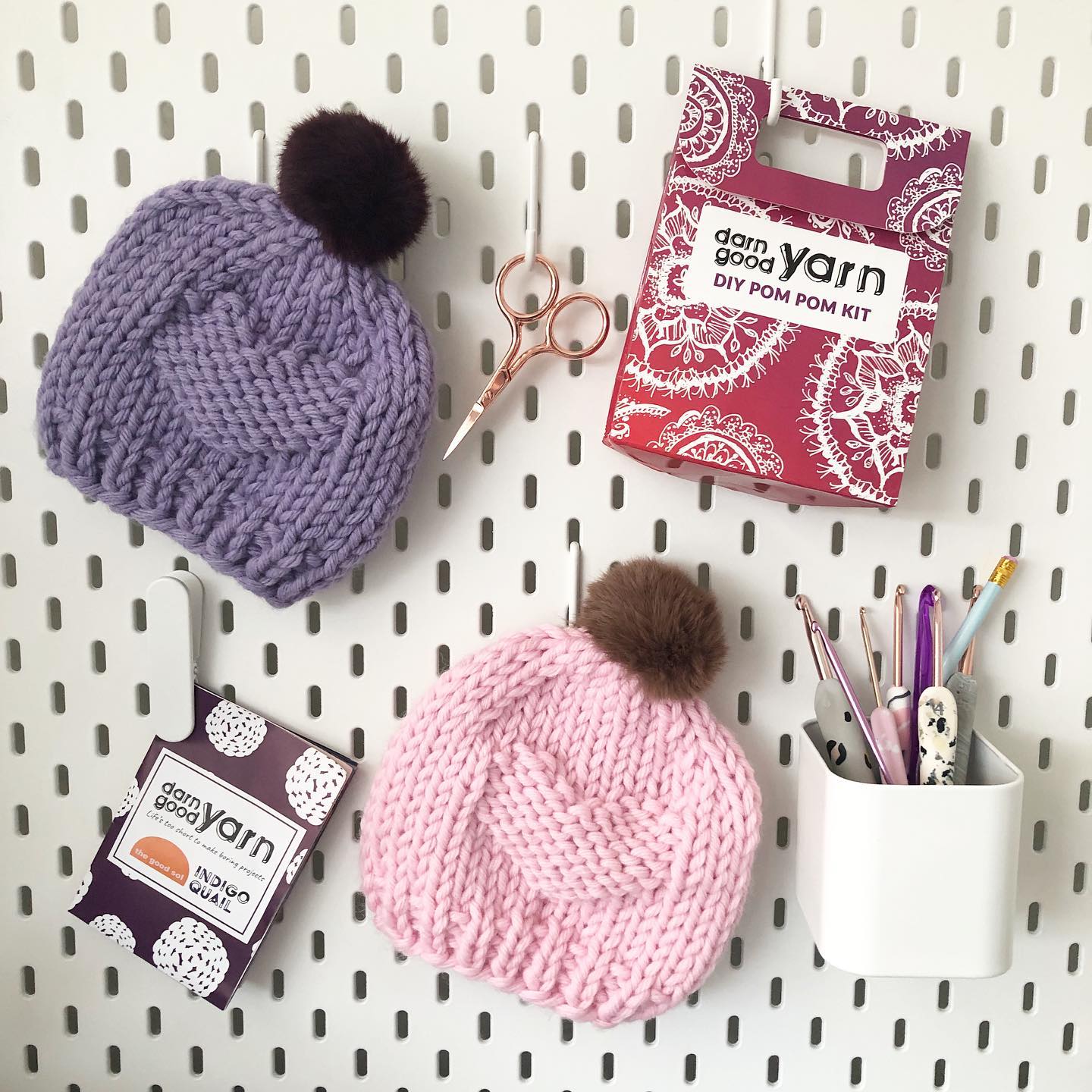 lifestyle image of pom pom kit, completed pom poms, and notions hanging in front of a white peg board.