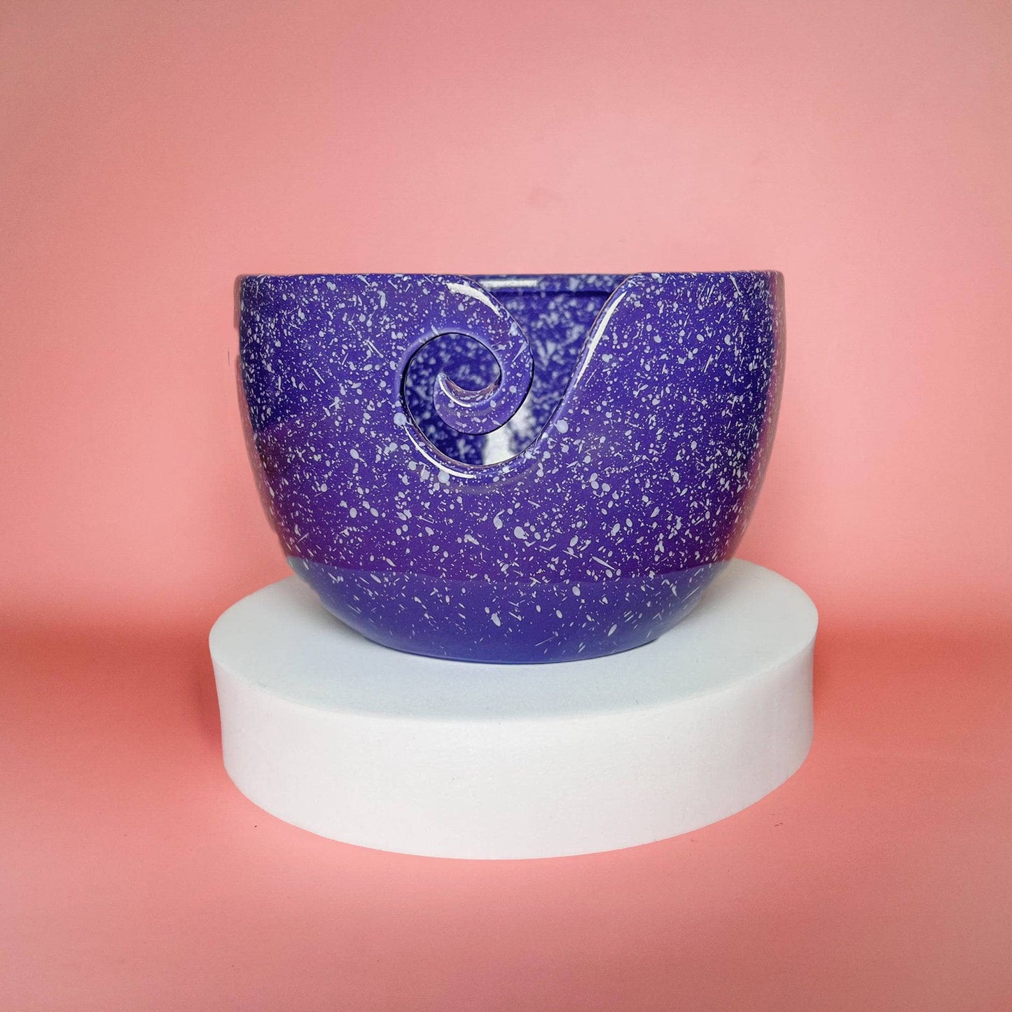 Purple yarn bowl with white speckles all over sitting on a round white pedestal in front of a pink background.