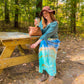 Young woman with tie dye dress, blue green long sleeve top, brown hat, leather boots sitting at a wooden picnic table in the woods and drinking coffee and smiling gently at the camera.
