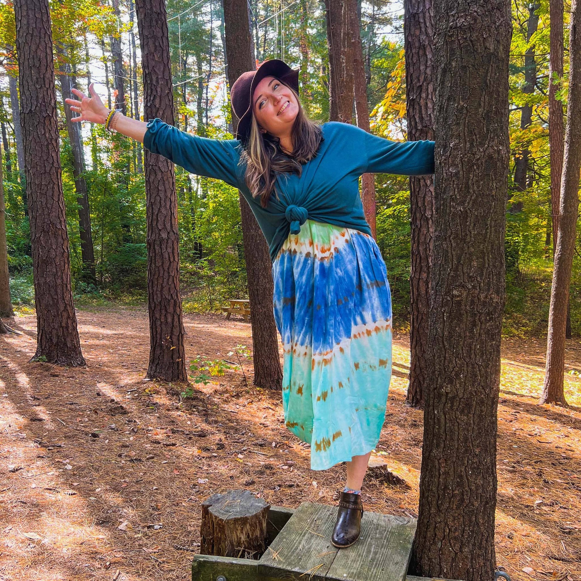 Young woman in tie dye dress, long sleeve blue green top and brown hat standing in woods and holding onto tree and leaning to one side smiling kindly at the camera.