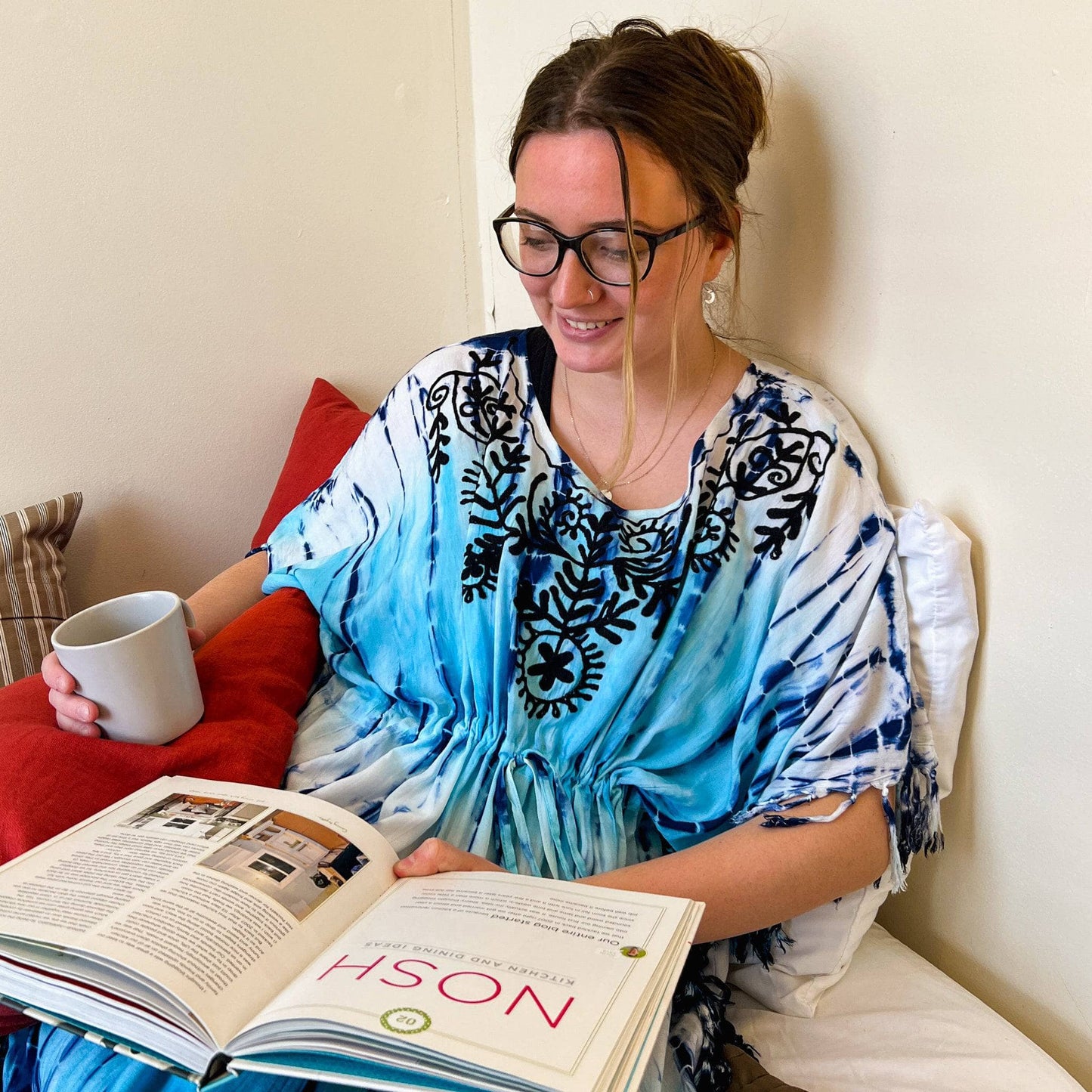 Woman wearing a blue tie dye tunic sitting in bed, reading a book.