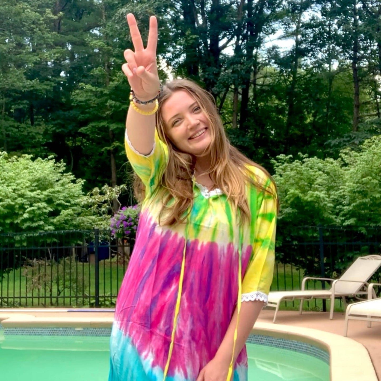 Model giving a peace sign wearing a brilliant tie dye cover up by the pool.